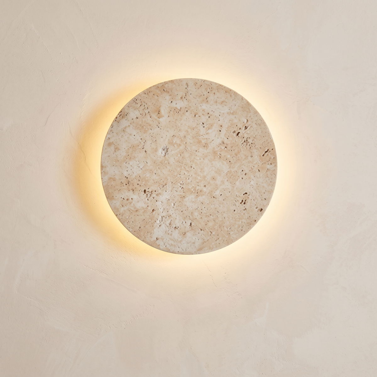 Matera round stone wall light with a warm halo glowing from behind the disc on a limewash wall