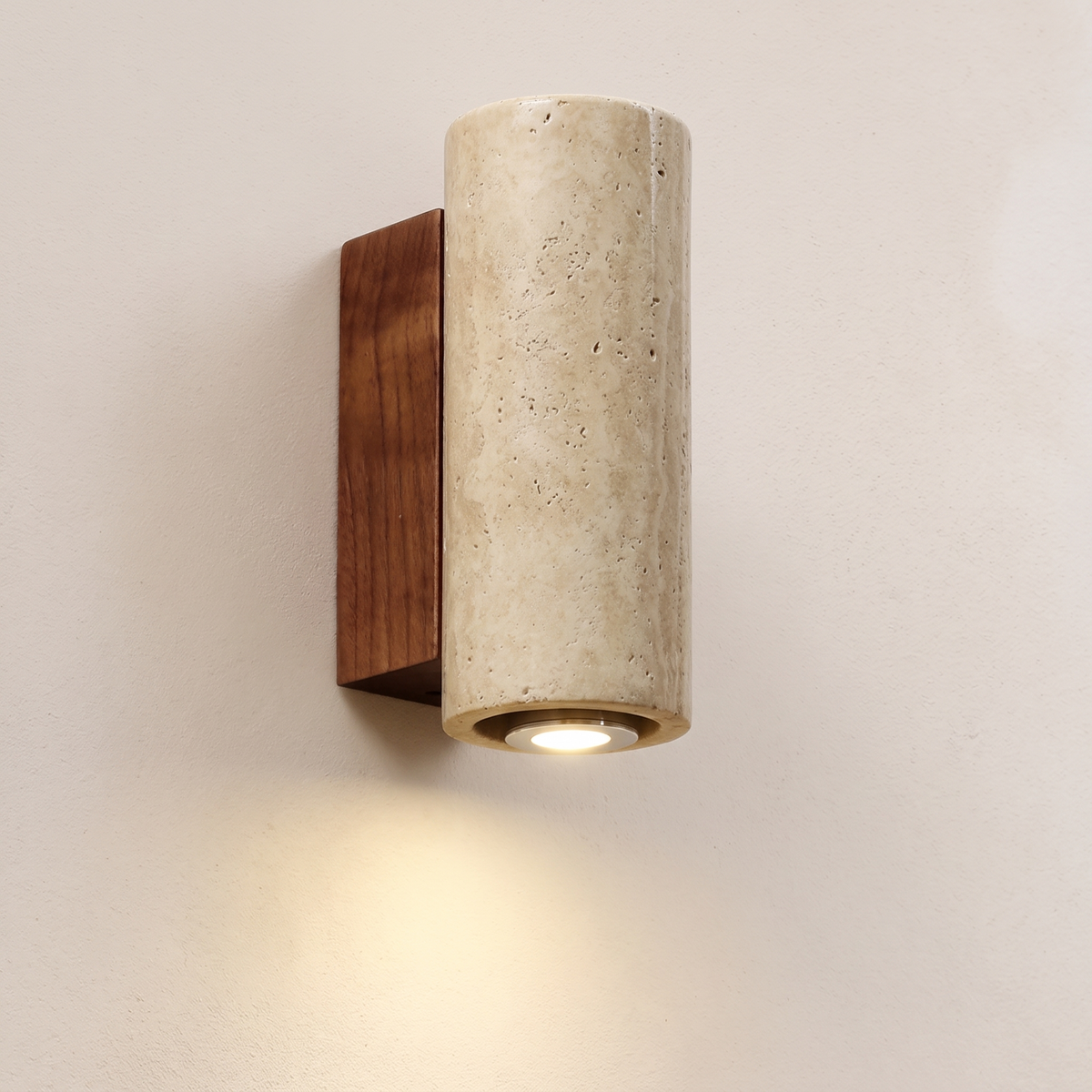 Tropea wall light, a round travertine cylinder on a walnut backplate, lit against a limewash wall